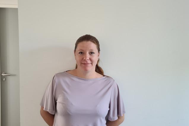 Frau mit langen Haaren steht l&auml;chelnd an einer Wand in einem hellen Raum, neben einer geschlossenen T&uuml;r und einem Feuerl&ouml;scher.