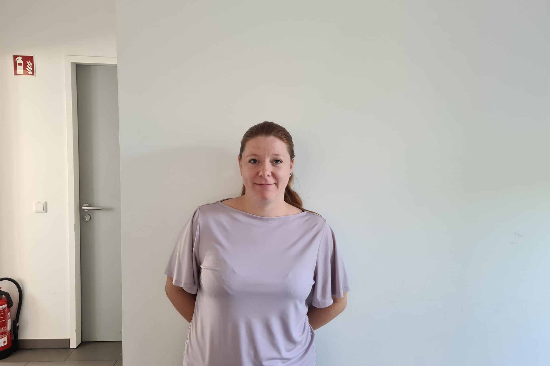 Frau mit langen Haaren steht lächelnd an einer Wand in einem hellen Raum, neben einer geschlossenen Tür und einem Feuerlöscher.