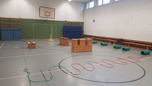 Leere Turnhalle mit zwei Holzkästen, einem Ständer und übereinandergelegten Matten. Bunte Seile liegen auf dem Boden.