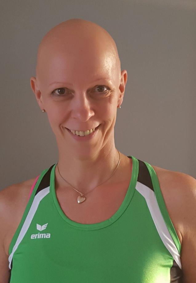 Frau mit Glatze trägt ein grünes Sporttop und lächelt in einmittleres Licht vor einer neutralen Wand.