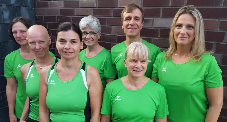 Gruppe von sieben Personen in grünen Sporttrikots, stehend vor einer Mauer, freundlich lächelnd.