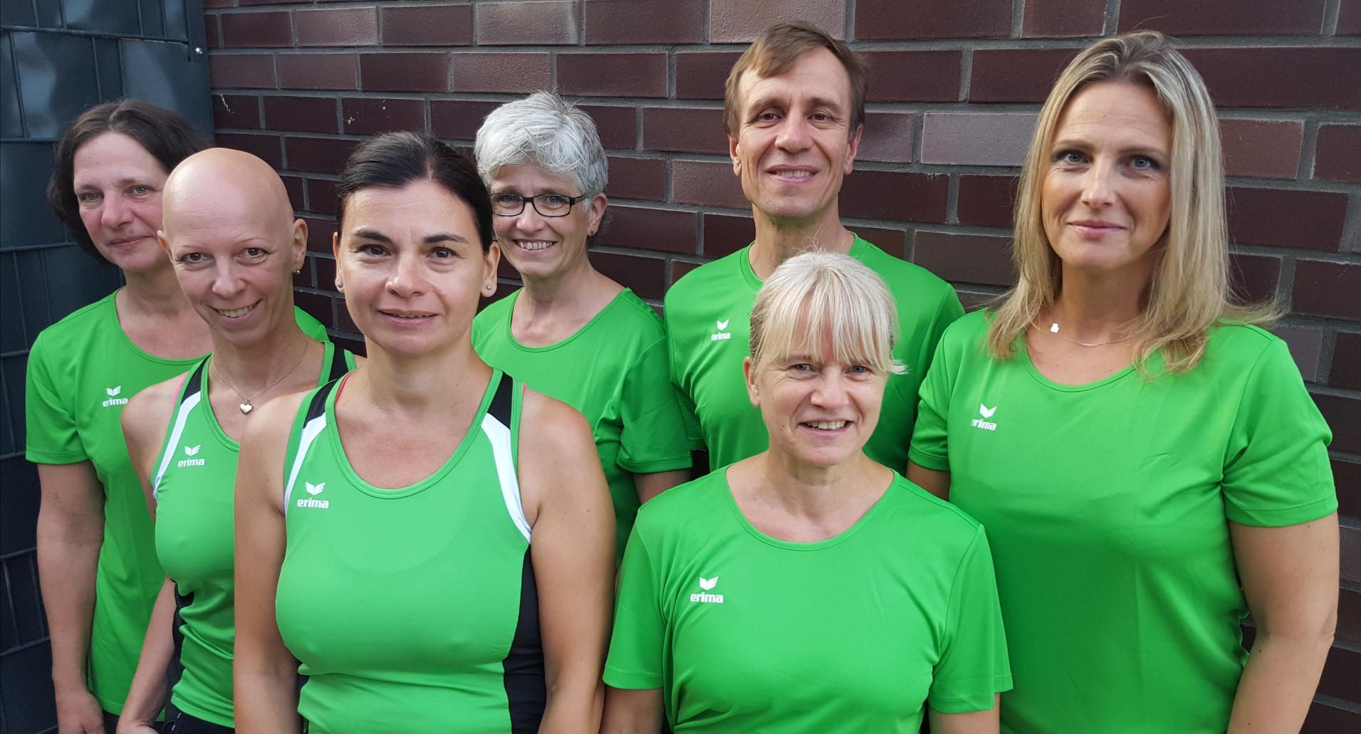 Gruppe von sieben Personen in grünen Sporttrikots, stehend vor einer Mauer, freundlich lächelnd.