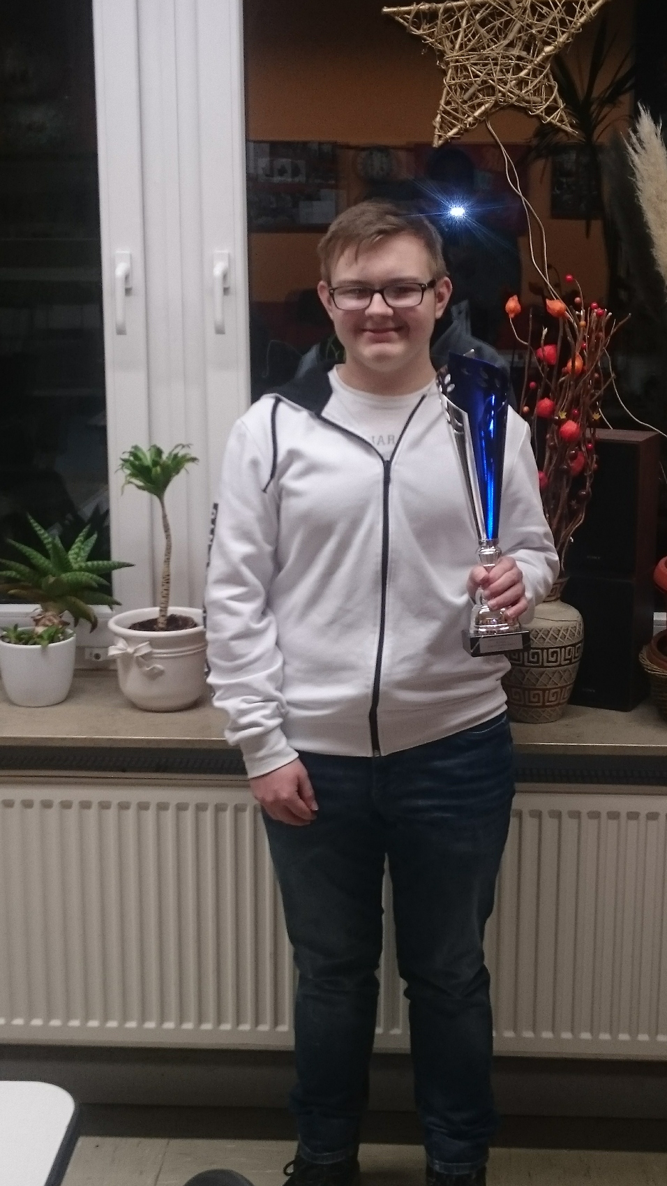 Junger Mann in weißem Hoodie hält eine blaue Trophäe vor einem Fenster mit Pflanzen und Dekoration im Hintergrund.
