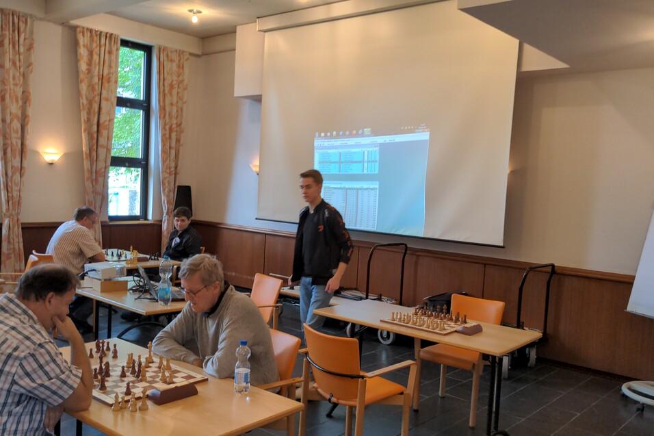 Schachspieler analysieren eine Partie in einem Raum mit einem Projektor und mehreren Schachbrettern auf Tischen.