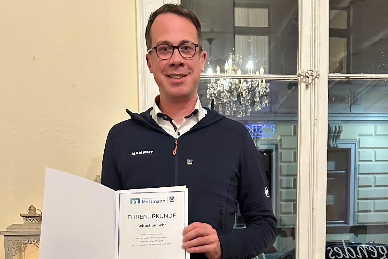 Mann h&auml;lt eine Ehrenurkunde in der Hand, steht vor einem Fenster mit dekorativen Elementen im Hintergrund.