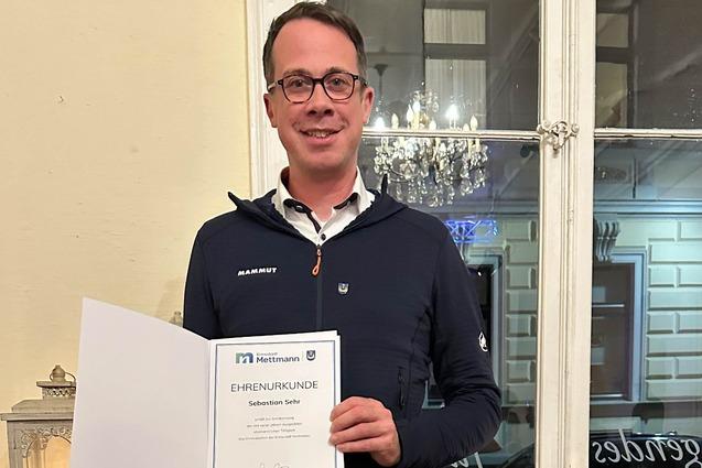 Mann hält eine Ehrenurkunde in der Hand, steht vor einem Fenster mit dekorativen Elementen im Hintergrund.