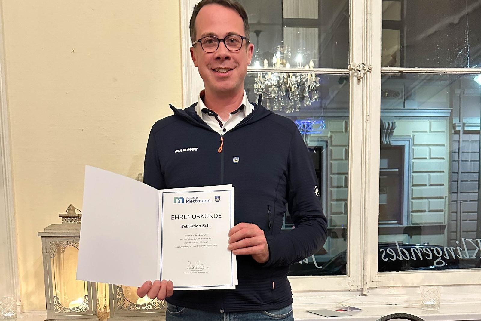 Mann hält eine Ehrenurkunde in der Hand, steht vor einem Fenster mit dekorativen Elementen im Hintergrund.