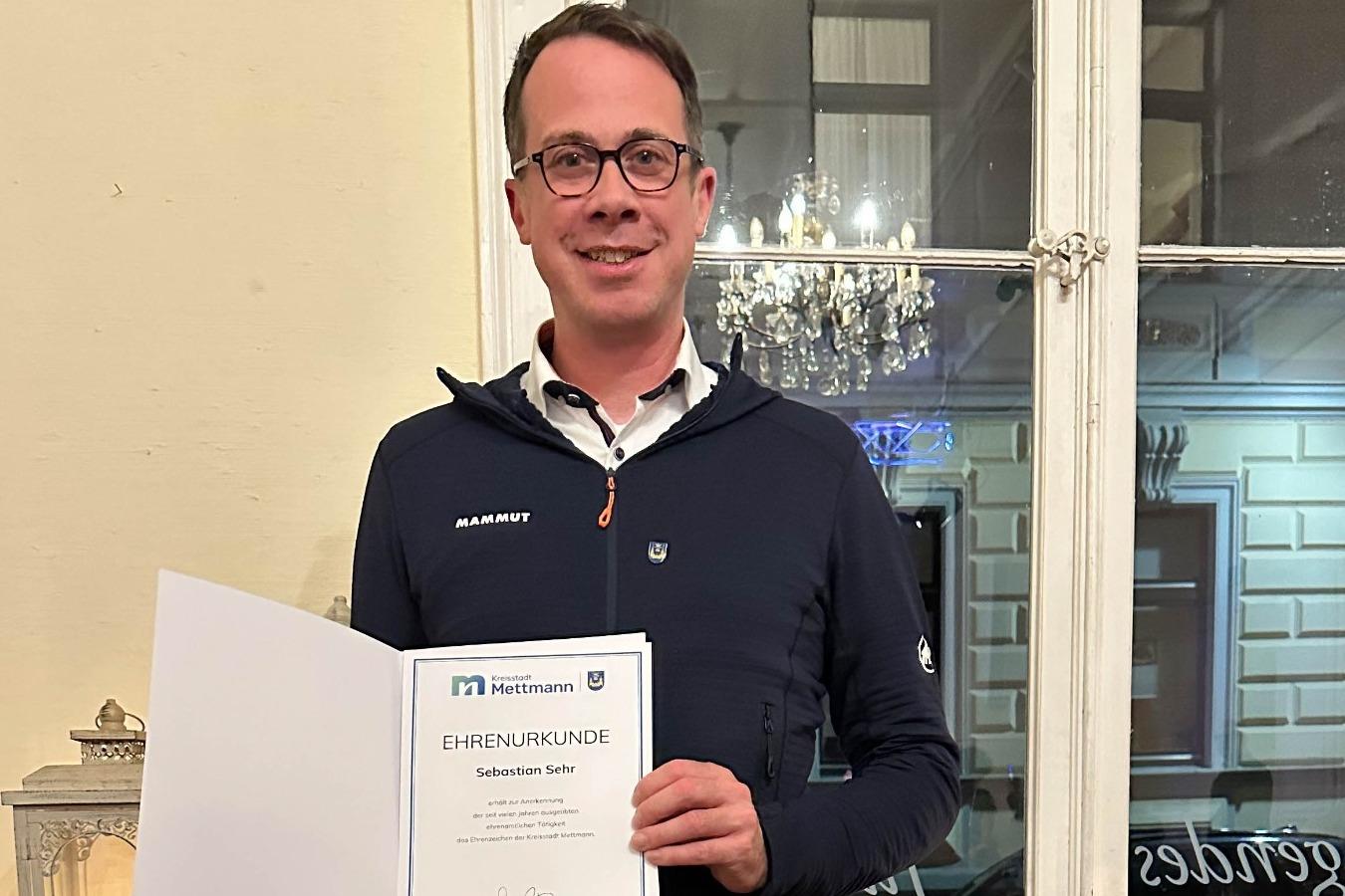 Mann h&auml;lt eine Ehrenurkunde in der Hand, steht vor einem Fenster mit dekorativen Elementen im Hintergrund.