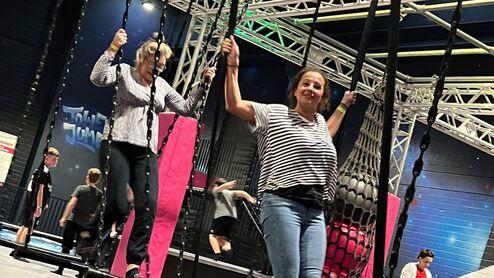 Zwei Frauen schwingen an Seilen in einem Trampolinpark, während im Hintergrund weitere Personen aktiv sind.