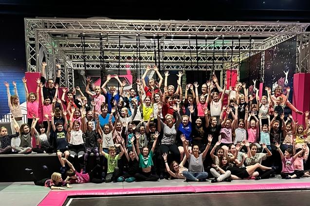 Gruppierung von Kindern und Jugendlichen in bunten T-Shirts, die fröhlich die Hände heben, vor einem Trampolinpark.