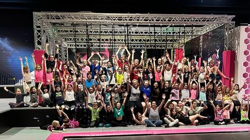 Gruppierung von Kindern und Jugendlichen in bunten T-Shirts, die fröhlich die Hände heben, vor einem Trampolinpark.