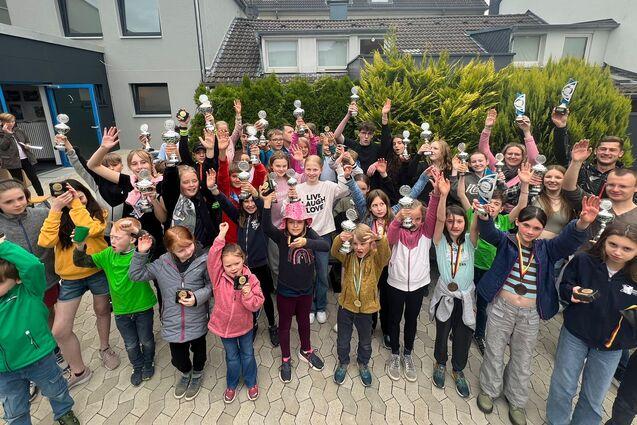 Gruppe von Kindern und Jugendlichen hält Trophäen und Medaillen hoch, umgeben von einem Wohnhaus und grünen Pflanzen.