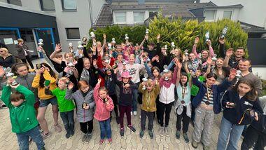 Gruppe von Kindern und Jugendlichen hält Trophäen und Medaillen hoch, umgeben von einem Wohnhaus und grünen Pflanzen.