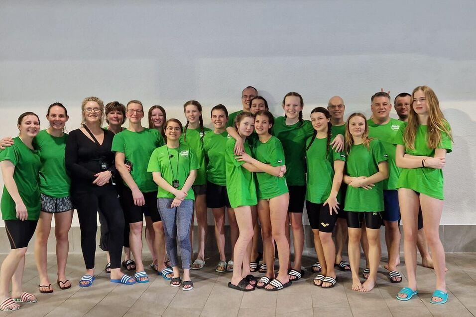Gruppe von Schwimmern in grünen T-Shirts posiert zusammen in einem Schwimmbad, Hintergrunde hell und neutral.