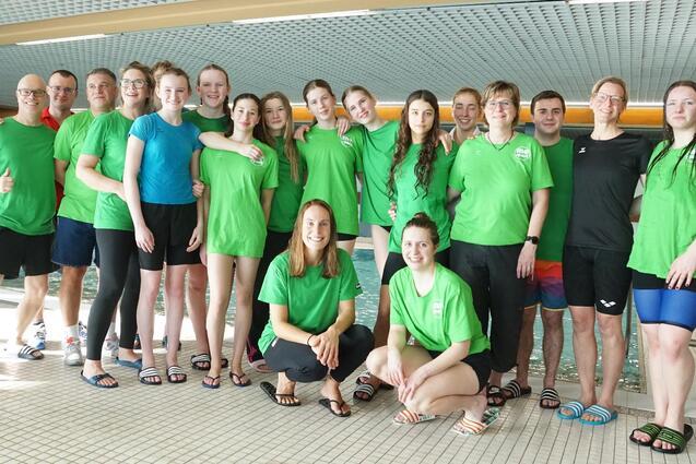 Gruppe von Schwimmern in grünen T-Shirts, posiert am Rand eines Schwimmbeckens in einem Hallenbad.