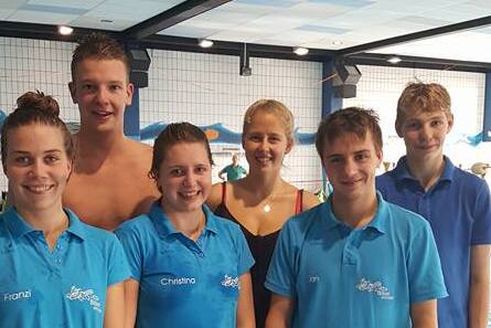 Sechs Schwimmer in blauen Jerseys stehen lächelnd am Beckenrand eines Schwimmbades.