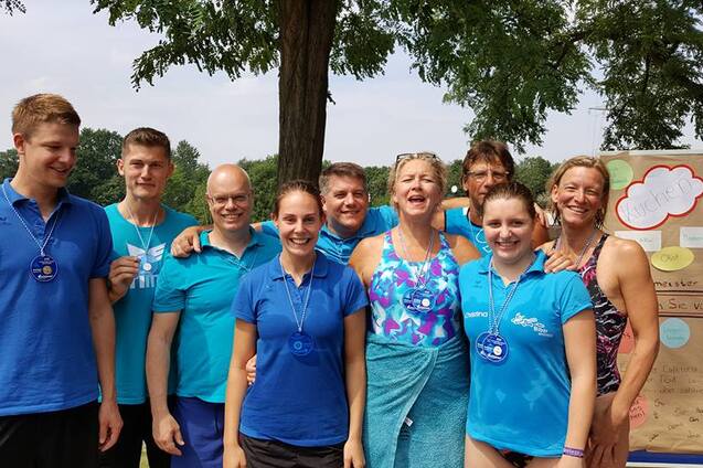 Gruppe von Menschen in blauen Shirts mit Medaillen, lächelnd vor einem Informationsposter im Freien.