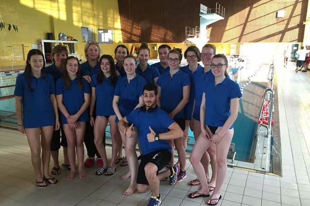 Gruppenfoto von Schwimmern und Trainern in blauen T-Shirts neben einem Schwimmbecken im Hallenbad.