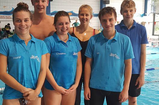 Sechs Schwimmer in blauen Jerseys stehen lächelnd am Beckenrand eines Schwimmbades.