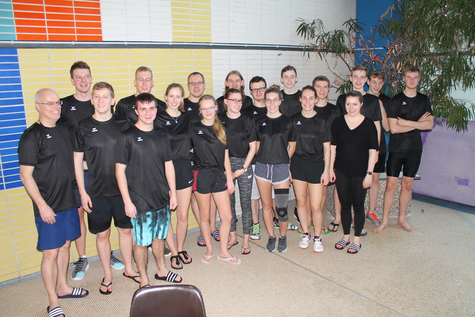 Gruppe von 17 sportlich gekleideten Jugendlichen und Erwachsenen in einem Hallenbad, umgeben von bunten Wänden.
