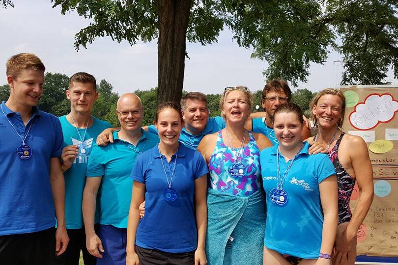 Gruppe von Menschen in blauen Shirts mit Medaillen, lächelnd vor einem Informationsposter im Freien.