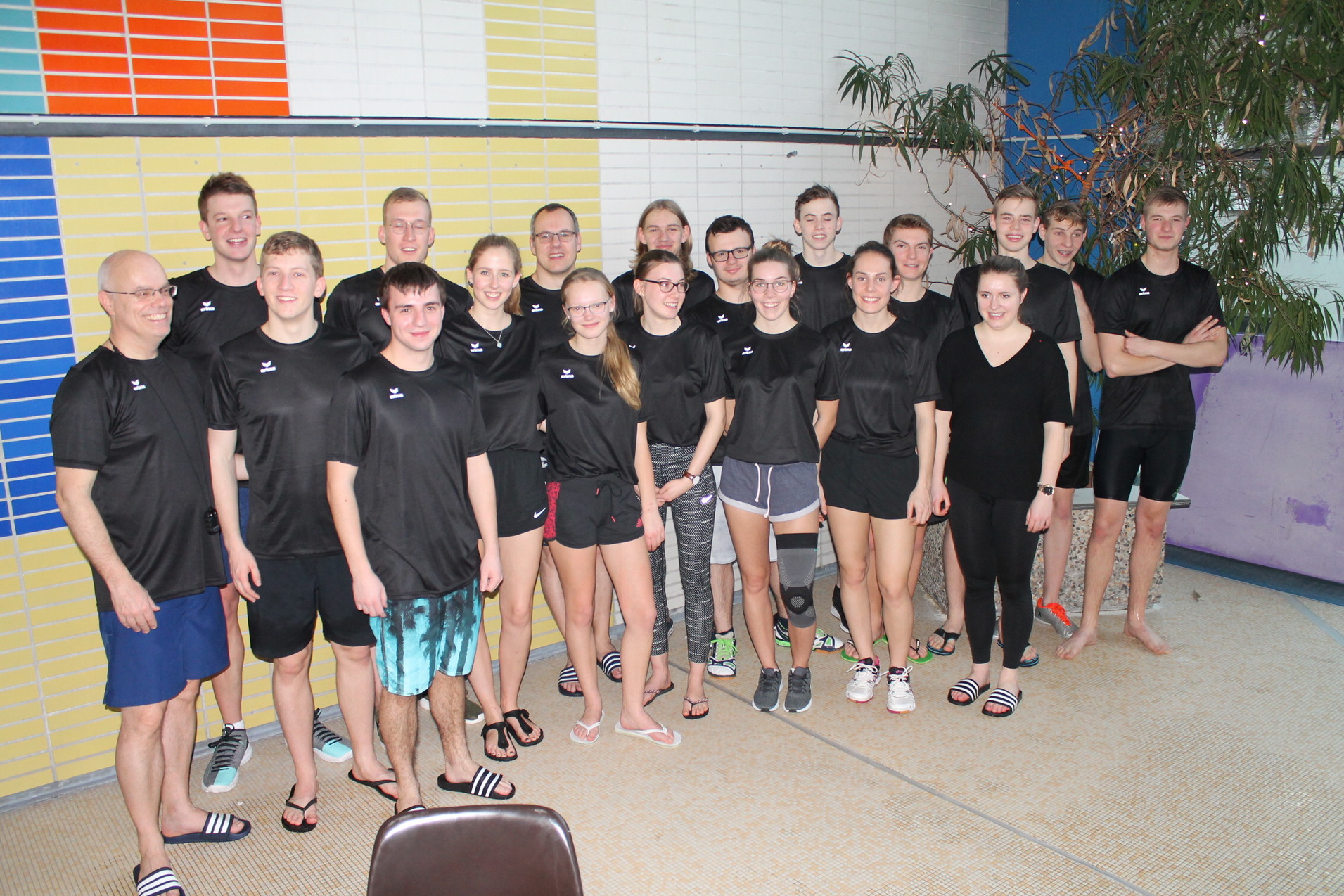 Gruppe von 17 sportlich gekleideten Jugendlichen und Erwachsenen in einem Hallenbad, umgeben von bunten Wänden.