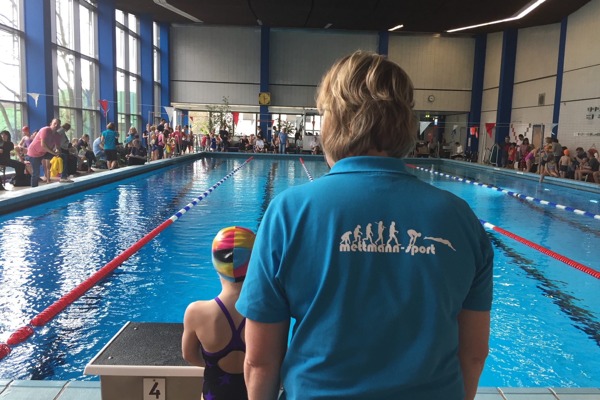 Eine Person in Blauen Shirt steht neben einem Schwimmbecken, ein Kind in Badekleidung mit bunter Schwimmhaube ist bereit zu starten.