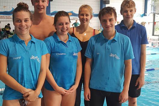 Sechs Schwimmer in blauen Jerseys stehen lächelnd am Beckenrand eines Schwimmbades.