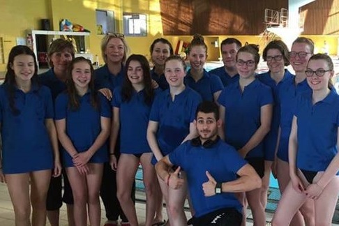Gruppenfoto von Schwimmern und Trainern in blauen T-Shirts neben einem Schwimmbecken im Hallenbad.