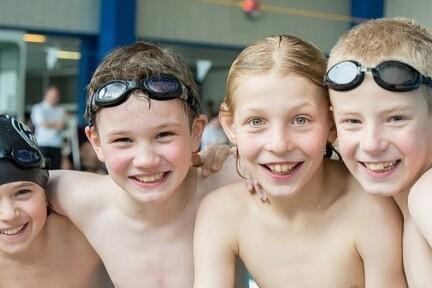 Gruppe von vier fröhlichen Schwimmkindern ohne T-Shirt, am Beckenrand, mit Schwimmbrillen.