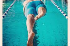 Sportler springt in ein Schwimmbecken, während er vom Startblock in blauer Badehose abspringt.