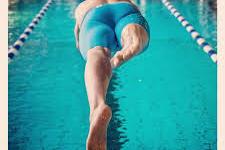 Sportler springt in ein Schwimmbecken, während er vom Startblock in blauer Badehose abspringt.