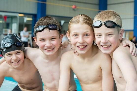 Gruppe von vier fröhlichen Schwimmkindern ohne T-Shirt, am Beckenrand, mit Schwimmbrillen.