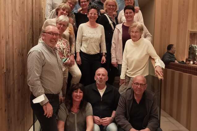 Gruppenselfie von 24 Personen in einem modernen Innenraum auf einer Treppe mit Holzverkleidung.