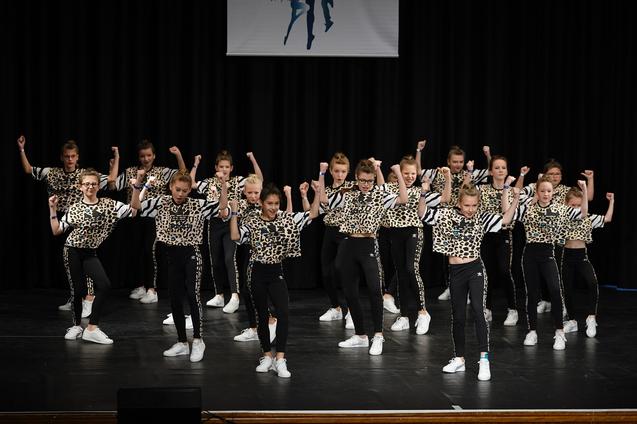 Gruppe von tanzenden Mädchen in Leoparden-Outfits auf einer Bühne, schwarzer Hintergrund und schwaches Licht.