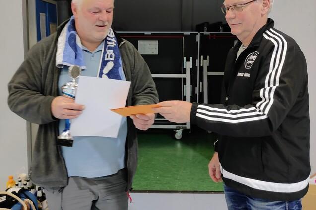Zwei Männer in einer Sporthalle überreichen sich eine Urkunde und einen Pokal. Der Mann links trägt ein Handtuch um den Hals.