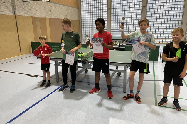 Vier Kinder stehen an einem Tischtennistisch, halten Pokale und Urkunden nach einem Wettbewerb.