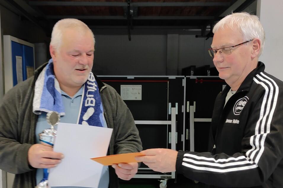 Zwei Männer in einer Sporthalle überreichen sich eine Urkunde und einen Pokal. Der Mann links trägt ein Handtuch um den Hals.