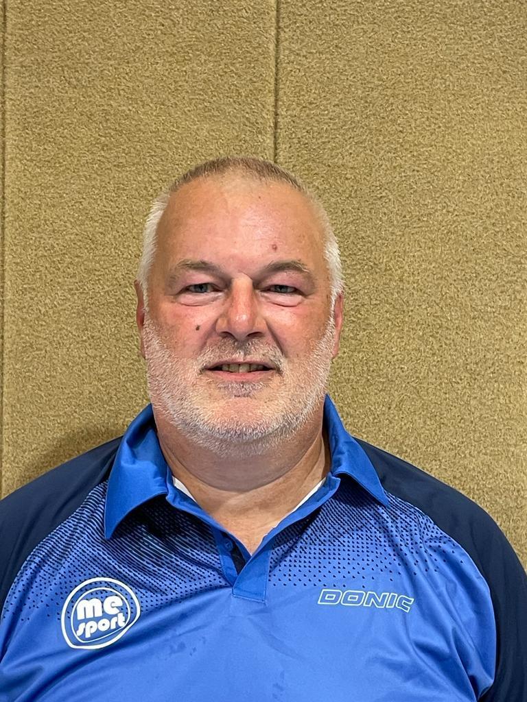 Mann mit kurzen grauen Haaren und Bart, trägt ein blaues Sporthemd, steht vor einem beige Wandhintergrund.