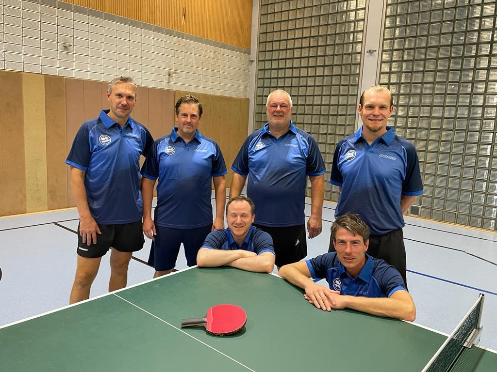 Sechs Tischtennisspieler in blauen Trikots posieren vor einem Tischtennistisch in einer Sporthalle.