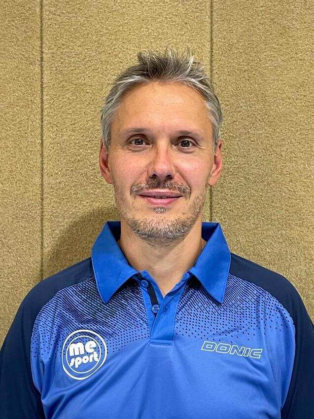 Lächelnder Mann mit kurzem, grauem Haar in einem blauen Polo-Shirt vor einer hellbraunen Wand.