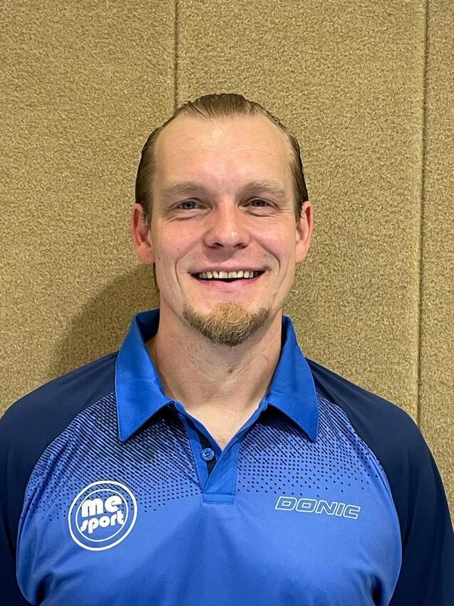 Portrait eines lächelnden Mannes in einem blauen Sporthemd mit Logo, vor einer beige Wand.