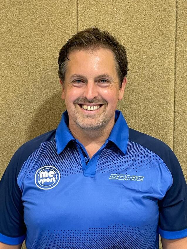 Ein lächelnder Mann trägt ein blaues Polo-Shirt mit Markenlogo vor einer hellbraunen Wand.