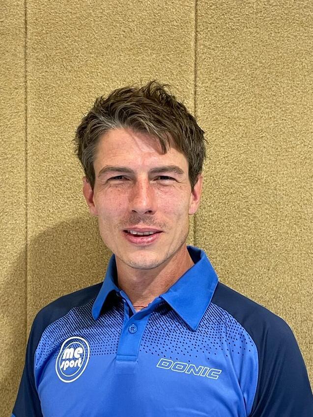 Porträt eines Mannes mit kurzem, geföhnter Haar, in einem blauen Shirt mit dem Logo „DONIC“ vor einer neutralen Wand.