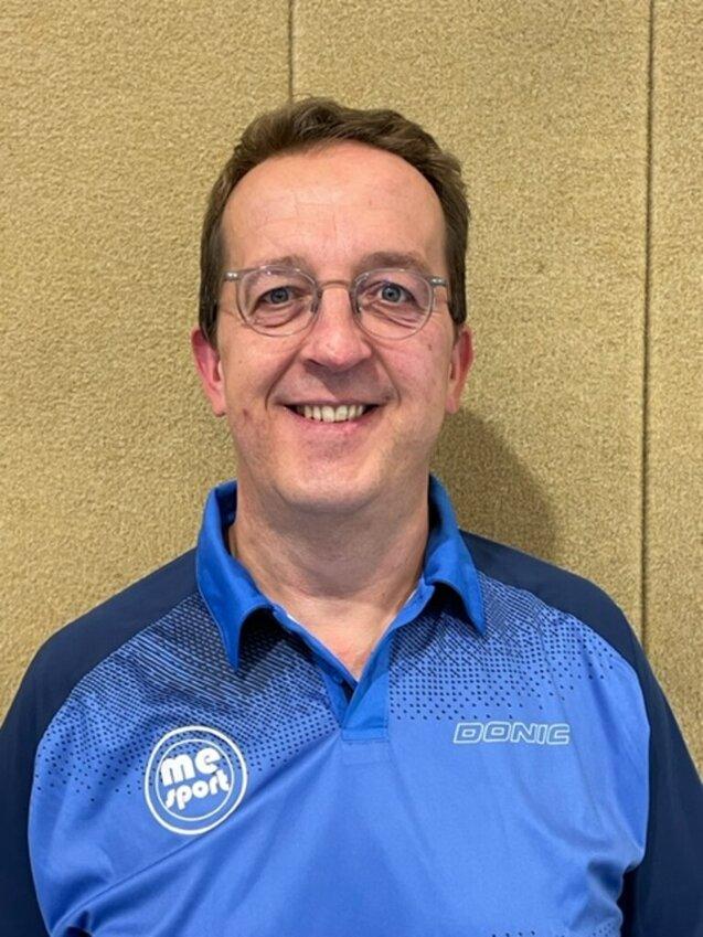 Lächelnder Mann mit Brille in blauem Sportshirt vor einer beige Wand.