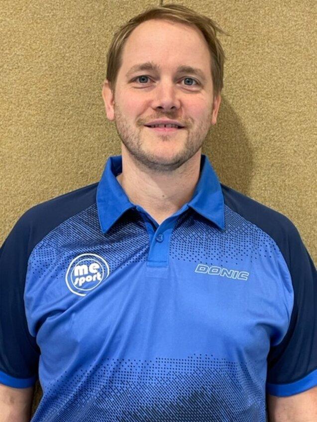 Porträt eines Mannes in blauer Sportkleidung mit Logo, vor einer neutralen, hellen Wand.