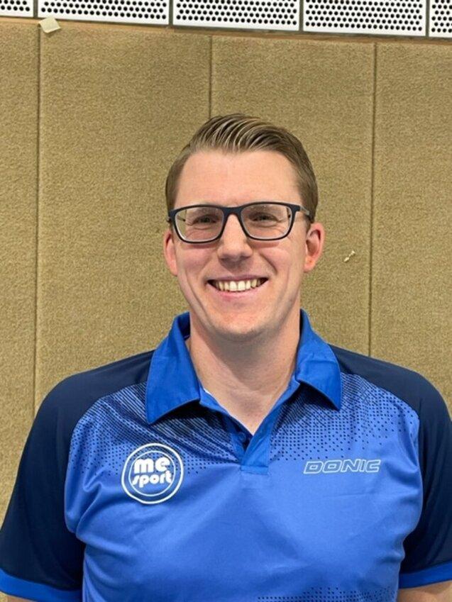 Lächelnder Mann mit Brille, in blauem Sportshirt mit Logo, vor einer beige Wand.