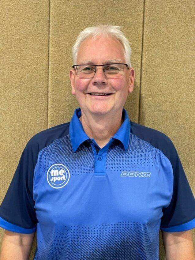 Mann mit Brille und breitem Lächeln trägt ein blaues Sportshirt vor einer schlichten Wand.