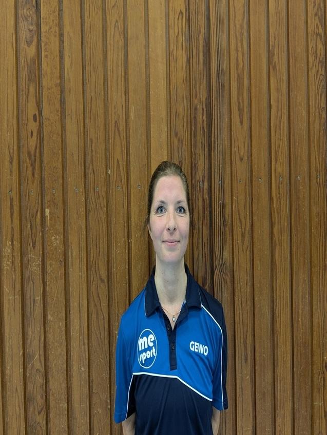 Portrait einer Frau mit blauen Augen in einem blauen Sportshirt vor einer Holzpaneelenwand.