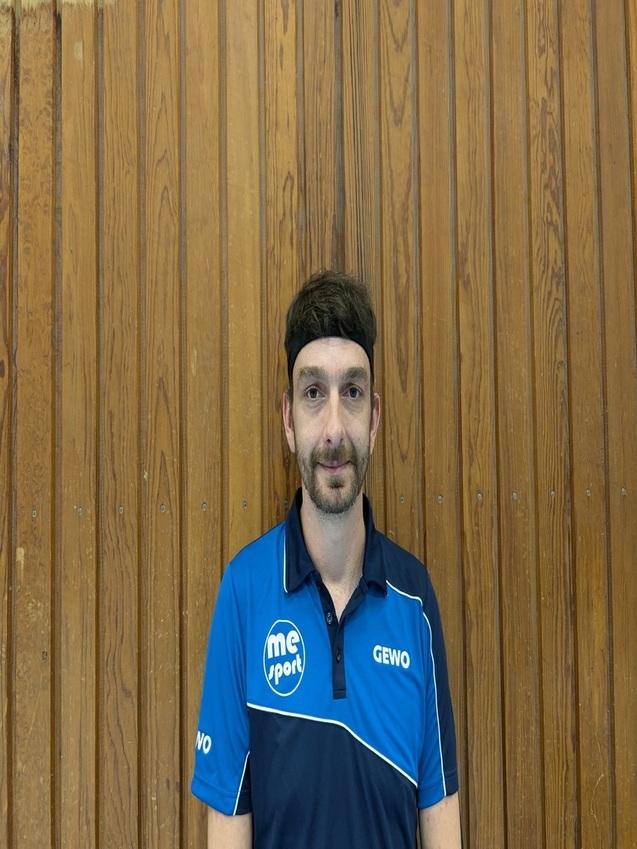 Mann mit Stirnband und blauem Sporttrikot steht vor einer Holzwand in einer Sporthalle.